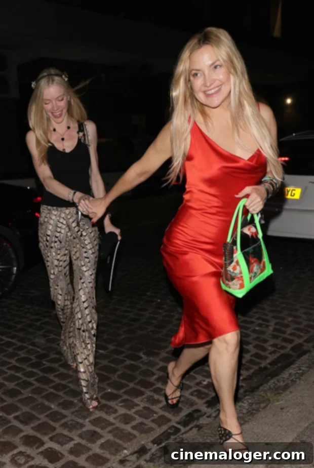 Red Hot Sisterhood: Kate Hudson & Ex's Wife's Glamorous Night Out 2 Kate Hudson and Elle Evans smile and hold hands as they walk out in London at night. Kate wears a red silky midi dress and black strappy heels, carrying a neon green bag. Elle wears snakeskin print pants and a black strapless top.